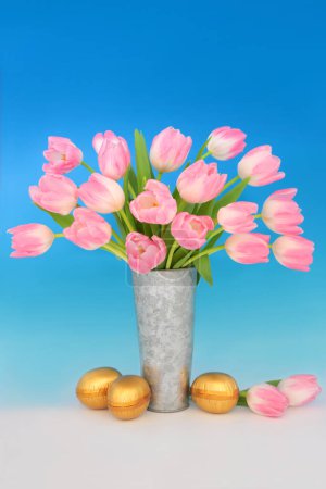 Photo for Gold Easter eggs and pink tulip flower bouquet arrangement in metal vase on gradient blue background. Luxury sweet food floral spring nature design for the holiday season. - Royalty Free Image