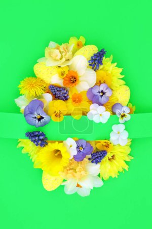 Easter egg concept shape with yellow blue white Spring flowers and decorated eggs on green background. Happy Easter composition for the festive season with copy space.