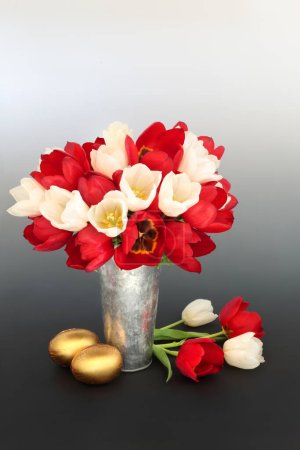 Gold chocolate Easter eggs and red tulip flowers in a vase on gradient black white background. Spring floral, festive luxury gifts for the holiday season.