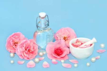 Rose flower perfume in heart shaped bottle with pearls, flowers and pink petals on blue. Natural fragrant floral  product, gift for Valentines Day, birthday, anniversary or Mothers day.