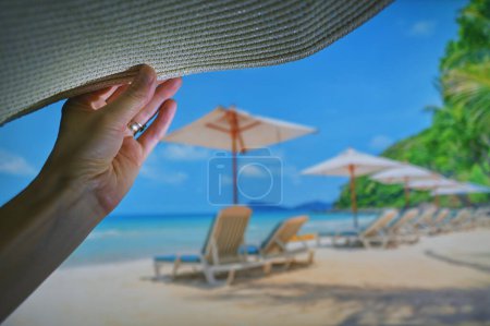 Téléchargez les photos : Grand angle de femme se détendre sur la plage tenant chapeau de soleil et la mer en arrière-plan - en image libre de droit