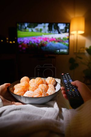 Téléchargez les photos : Gros plan femme manger tangrine et regarder la télévision sur lit - en image libre de droit