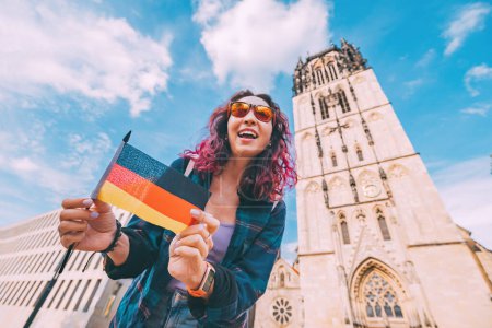 Foto de Una joven feliz turista o estudiante con una bandera alemana en el casco antiguo o Altstadt en Munster con campanario de la iglesia en el fondo. Estudiar el idioma en el extranjero y viajar concepto - Imagen libre de derechos