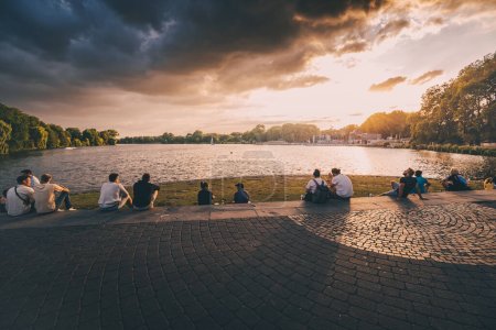Foto de 25 de julio de 2022, Munster, Alemania: Personas descansando en el parque de la ciudad de Munster y observando el atardecer sobre el lago Aasee - Imagen libre de derechos