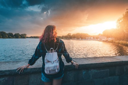 Foto de Una estudiante con una mochila se encuentra cerca del terraplén del lago Aasee en Munster y disfruta de una impresionante vista al atardecer - Imagen libre de derechos