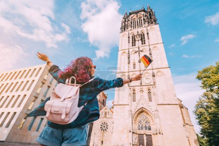 Foto de Una joven feliz turista o estudiante con una bandera alemana en el casco antiguo o Altstadt en Munster con campanario de la iglesia en el fondo. Estudiar el idioma en el extranjero y viajar concepto - Imagen libre de derechos