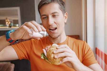 Foto de Un hombre con una cara alegre derrama un exceso de sal en su hamburguesa en un restaurante. Riesgo de enfermedades cardiovasculares y concepto de adicción - Imagen libre de derechos