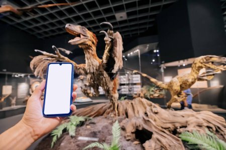 Foto de 26 de julio de 2022, Museo de Historia Natural de Munster, Alemania: Mano de visitante con pantalla de teléfono inteligente en blanco en la Exposición del aterrador dinosaurio emplumado deinonychus - Imagen libre de derechos