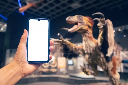 Foto de 26 de julio de 2022, Museo de Historia Natural de Munster, Alemania: Mano de visitante con pantalla de teléfono inteligente en blanco en la Exposición del aterrador dinosaurio emplumado deinonychus - Imagen libre de derechos