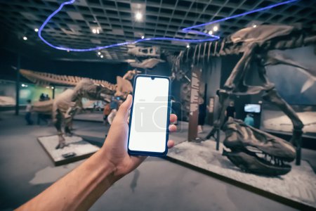 Foto de 26 de julio de 2022, Munster Natural History Museum, Alemania: Mano de visitante con pantalla de smartphone en blanco en la Exposición de esqueletos fósiles dinosaurios - Imagen libre de derechos