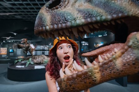 Foto de 26 julio 2022, Munster, Alemania: Divertida turista o estudiante paleontóloga mira con interés la mandíbula abierta con muchos dientes grandes de un dinosaurio en el Museo de Historia Natural - Imagen libre de derechos