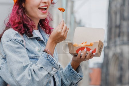Foto de 25 julio 2022, Munster, Alemania: Alegre chica feliz merienda en Nordsee fastfood con camarones en masa en la calle de la ciudad con una caja de papel para llevar - Imagen libre de derechos