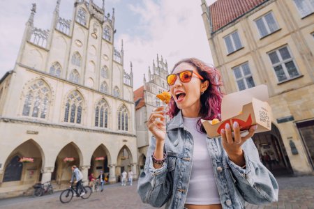 Foto de 25 julio 2022, Munster, Alemania: Alegre chica feliz merienda en Nordsee fastfood con camarones en masa en la calle de la ciudad con una caja de papel para llevar - Imagen libre de derechos