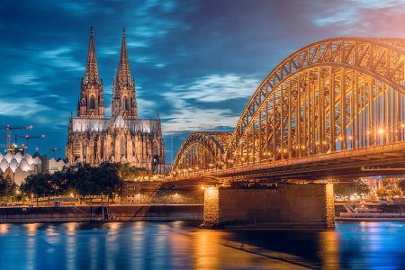 Téléchargez les photos : Vue panoramique du pont illuminé sur le Rhin, des trains et des touristes passant devant et de la cathédrale de Cologne le soir à l'heure bleue. - en image libre de droit