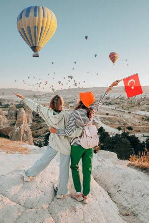 Foto de Amigas viajeras con bandera turca, abrazándose en un mirador y admirando la vista de los globos aerostáticos voladores en Capadocia - Imagen libre de derechos