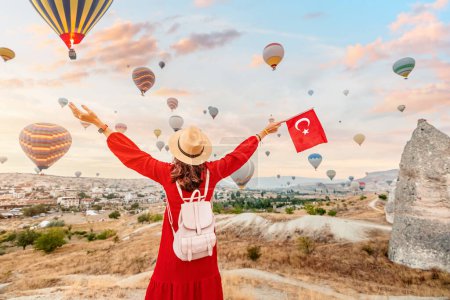 Téléchargez les photos : Joignez-vous à une jeune femme qui regarde les emblématiques montgolfières de Cappadoce, en Turquie, tout en arborant fièrement le drapeau turc. Une célébration de la beauté et de la culture du pays. - en image libre de droit