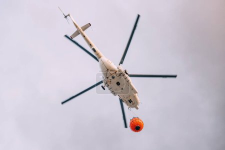 Foto de Un helicóptero de emergencia de bombero volando a la zona de accidentes con una cesta con agua contra el fondo del cielo - Imagen libre de derechos