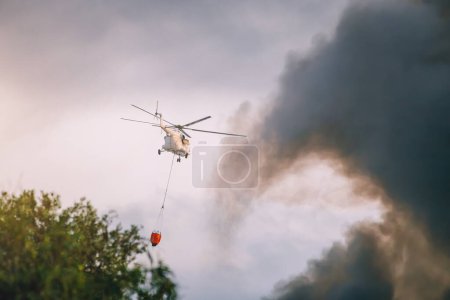 Foto de Un helicóptero de emergencia de bombero apaga un fuego y rocía agua de una canasta sobre una columna de humo negro sobre una ciudad o un incendio forestal - Imagen libre de derechos