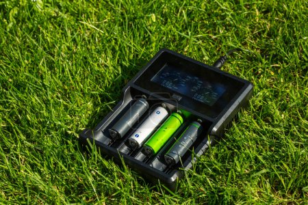 Téléchargez les photos : Chargeur de piles AA noir avec piles alcalines rechargeables sur l'herbe. La batterie se charge. - en image libre de droit