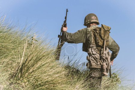 Téléchargez les photos : Reeneactor historique habillé comme soldat d'infanterie de la Seconde Guerre mondiale gravir une colline couverte d'herbe haute. Vue de l'arrière. - en image libre de droit