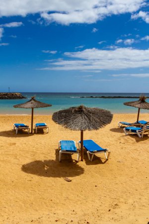 Ein leerer Strand in der Nebensaison mit Strohschirmen und blauen Sonnenliegen. Playa del Castillo in Caleta de Fuste, Fuerteventura, Kanarische Inseln, Spanien,