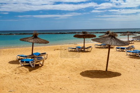 Une plage vide hors saison avec parasols en paille et chaises longues bleues. Playa del Castillo à Caleta de Fuste, Fuerteventura, Îles Canaries, Espagne,