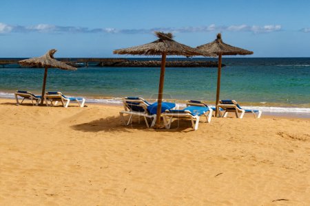 Una playa vacía en temporada baja con sombrillas de paja y tumbonas azules. Playa del Castillo en Caleta de Fuste, Fuerteventura, Islas Canarias, España.