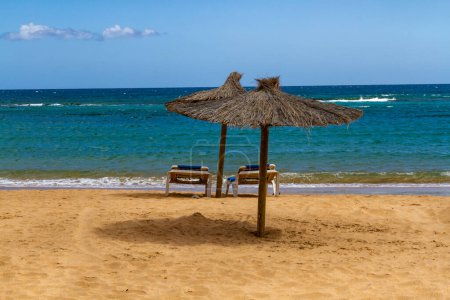 An empty beach in the off-season with straw umbrellas and blue sun lounger.  Playa del Castillo in  Caleta de Fuste, Fuerteventura, Canary Islands, Spain.