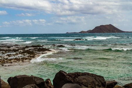 Photo for The rocky, volcanic coastline of the Atlantic Ocean near the port of Corralejo. Labos island in the background. Fuerteventura, Canary Islands, Spain - Royalty Free Image