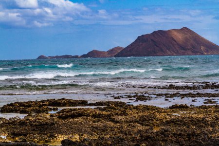 The rocky, volcanic coastline of the Atlantic Ocean near the port of Corralejo. Labos island in the background. Fuerteventura, Canary Islands, Spain