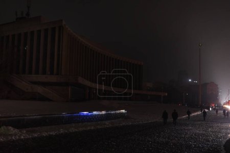 Foto de KYIV, UCRANIA - 16 de diciembre de 2022: Sala de conciertos Ucrania durante el apagón. Los transeúntes individuales se pueden ver en el pavimento cubierto de nieve por la noche. - Imagen libre de derechos