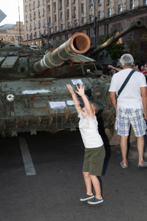Téléchargez les photos : KYIV, UKRAINE - 24 août 2023 : Un enfant tente d'atteindre le canon d'un char russe incendié lors d'une exposition de matériel militaire russe détruit - en image libre de droit
