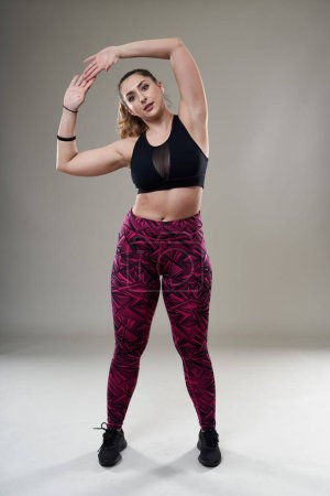 Photo for Confident plus size woman warming up and stretching before her workout, isolated on gray background - Royalty Free Image