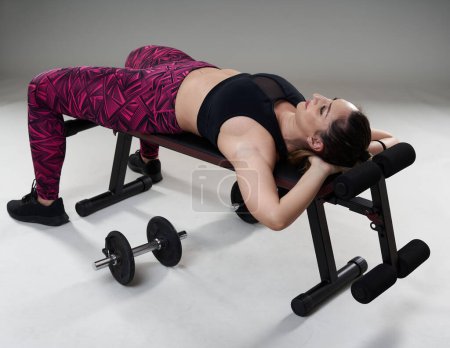 Foto de Mujer atractiva y segura de tamaño grande haciendo ejercicio con pesas en un banco de fitness, aislada sobre fondo gris - Imagen libre de derechos