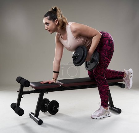 Photo for Confident attractive plus size woman working out with dumbbells on a fitness bench, isolated on gray background - Royalty Free Image