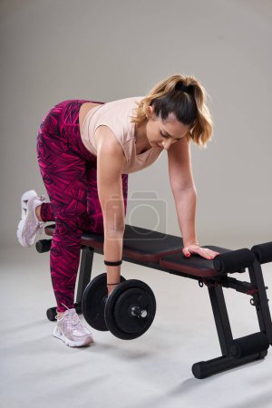 Photo for Confident attractive plus size woman working out with dumbbells on a fitness bench, isolated on gray background - Royalty Free Image