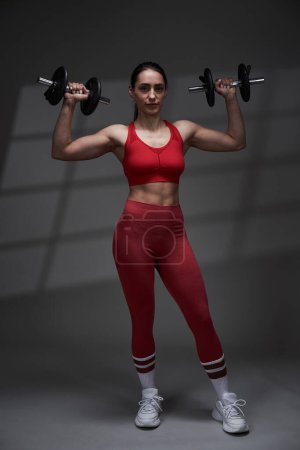 Téléchargez les photos : Athlétique jeune femme modèle de remise en forme en tenue de gymnastique rouge travailler, éclairage studio avec projection d'ombre sur le fond - en image libre de droit