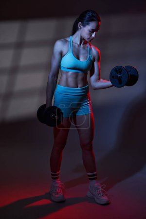 Téléchargez les photos : Modèle sportif de fitness jeune femme avec un corps parfait s'exerçant avec des haltères dans une lumière tonifiante bleu rouge dramatique avec projection de fenêtre sur le fond, travail en studio - en image libre de droit