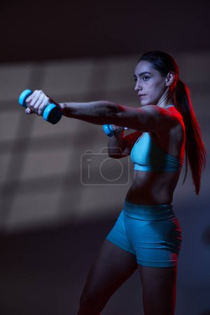 Photo for Athletic young woman fitness model with perfect body exercising with dumbbells in dramatic red blue toning light with window projection on background, studio work - Royalty Free Image