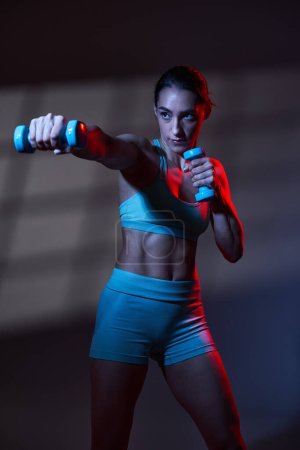 Téléchargez les photos : Modèle sportif de fitness jeune femme avec un corps parfait s'exerçant avec des haltères dans une lumière tonifiante bleu rouge dramatique avec projection de fenêtre sur le fond, travail en studio - en image libre de droit