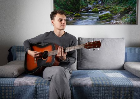 Foto de Young man playing an acoustic guitar on the sofa in the living room - Imagen libre de derechos