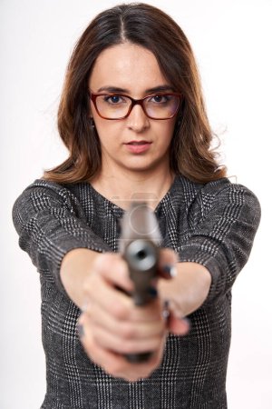 Photo for Businesswoman in reading glasses with serious expression pointing a gun at the camera, isolated on white background - Royalty Free Image