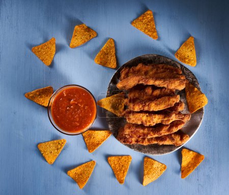 Foto de Chicken strips, croquettes with salsa sauce and nacho chips on a blue wooden board - Imagen libre de derechos