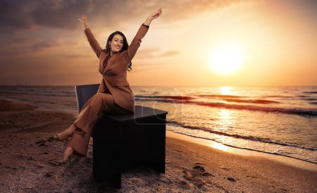 Téléchargez les photos : Businesswoman sitting on her desk placed on the ocean's shore at sunset, holiday concept - en image libre de droit