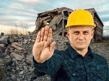 Téléchargez les photos : Engineer in hard hat making stop sign in front of ruined buildings - en image libre de droit