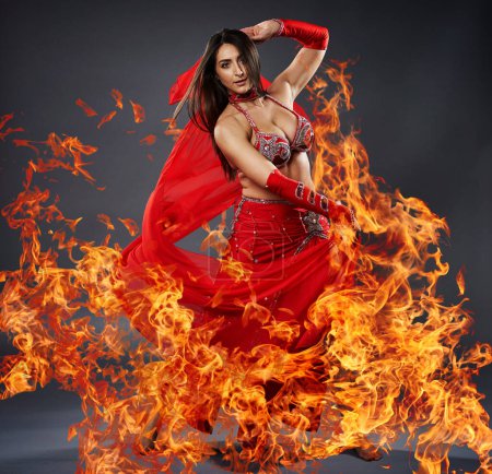 Téléchargez les photos : Arab woman belly dancer in red sparkling costume surrounded by flames dancing with her on gray background - en image libre de droit