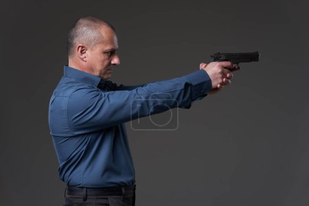 Photo for Mature businessman with a gun taking aim at the competition, gray background - Royalty Free Image