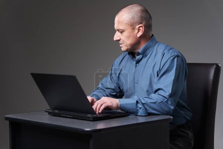 Téléchargez les photos : Mature businessman sitting at his desk working on laptop, on gray background - en image libre de droit