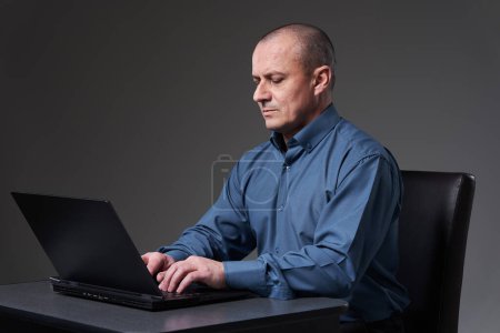 Photo for Mature businessman sitting at his desk working on laptop, on gray background - Royalty Free Image