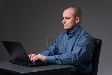 Foto de Mature businessman sitting at his desk working on laptop, on gray background - Imagen libre de derechos